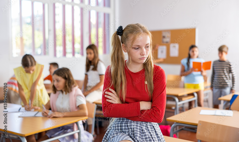 Upset tween girl sitting on table in schoolroom during recess on background with other pupils..