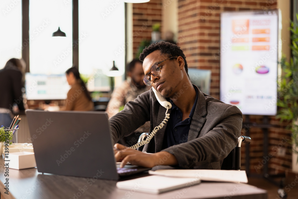Businessman with a disability attending a client call and typing on his laptop in brick wall workplace. African american entrepreneur in wheelchair using a landline phone and laptop in startup office. Businessman with a disability attending a client call and typing on his laptop in brick wall workplace. African american entrepreneur in wheelchair using a landline phone and laptop in startup office.