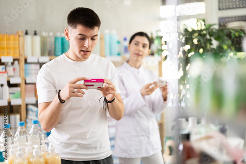 Young man buyer choosing box of paracetamol tablets with help of young woman pharmacist in pharmacy