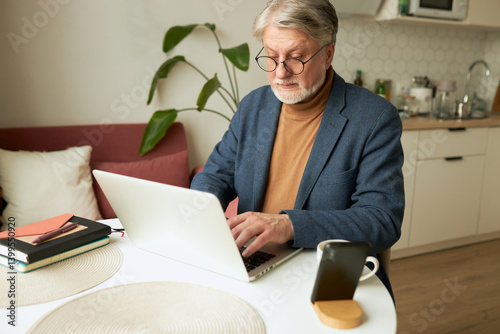 Side view of mature elegant businessman sitting at table in kitchen and working at laptop, finishing project. Senior male CEO in jacket checking e-mail from home at his desk with smartphone and cup