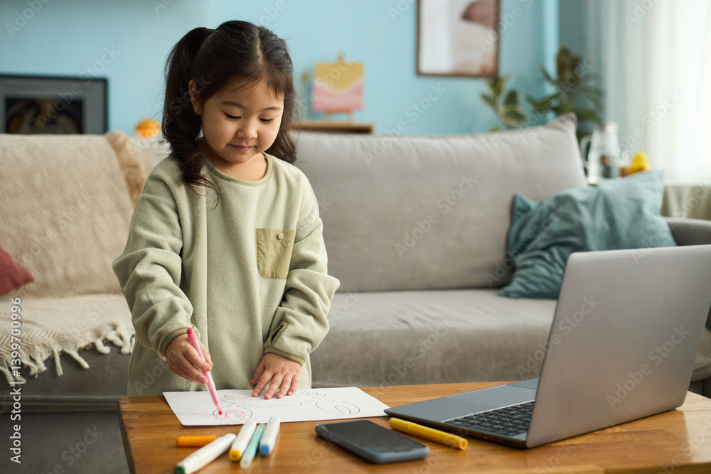 Young girl sitting at table drawing with markers while smiling, laptop and smartphone nearby on wooden table, home interior with sofa in background Young girl sitting at table drawing with markers while smiling, laptop and smartphone nearby on wooden table, home interior with sofa in background