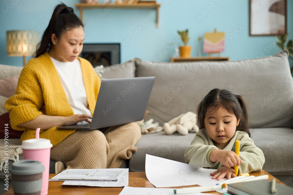 Asian mother working on laptop while young daughter engaged in drawing at living room table, illustrating daily life and productivity Asian mother working on laptop while young daughter engaged in drawing at living room table, illustrating daily life and productivity