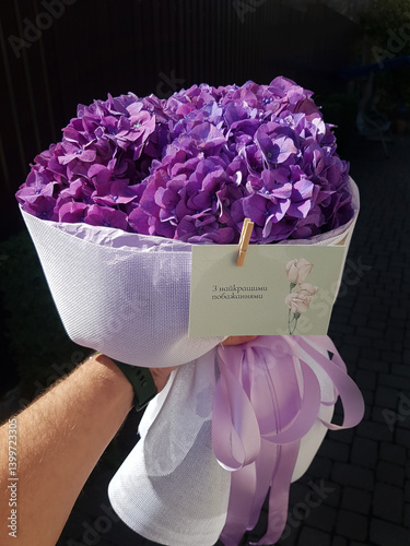 Image features a close-up of a bouquet of vibrant purple hydrangeas wrapped in a textured white fabric. The bouquet is held by a hand with light skin, wearing a dark green wristband.