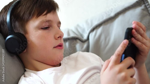 Teen boy watching short videos on phone lying on bed wearing black headphones