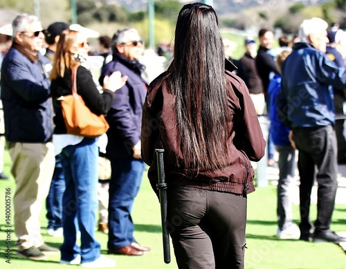 Vigilante de seguridad privada mujer con porra de espaldas protegiendo recinto al aire libre entre personas aficionadas de público
