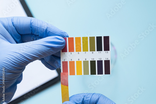 Hands holding ph test strip with color scale on blue background. Acidic pH environment.