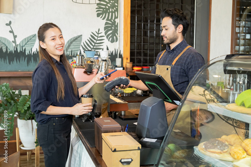 Young Indian baristas or cashier workers man holding a payment machine for scan, cashless technology and credit card payment concept. Customer using smartphone for payment to owner at cafe restaurant