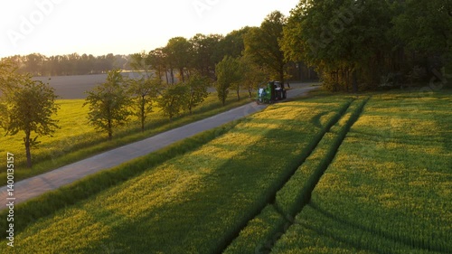 Drone shot of a tractor towing a header on a road during golden hour. The last rays of sunlight cast a warm glow over the rural landscape. Peaceful end to a farm day.