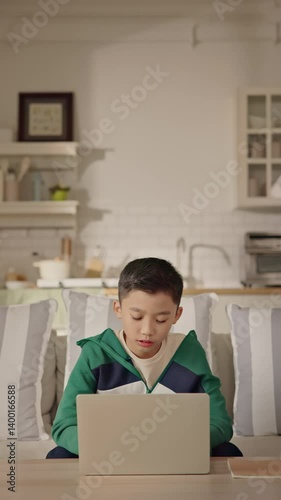 A young Asian boy sits comfortably at home using a laptop for gaming, studying, or browsing online. Modern childhood, digital education, balance of learning and leisure through technology. Gen Alpha.