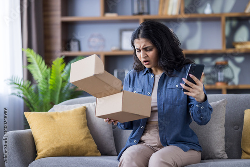 Worried young Indian woman sitting on the couch at home, holding a mobile phone and looking in shock at the parcel box she received in the mail
