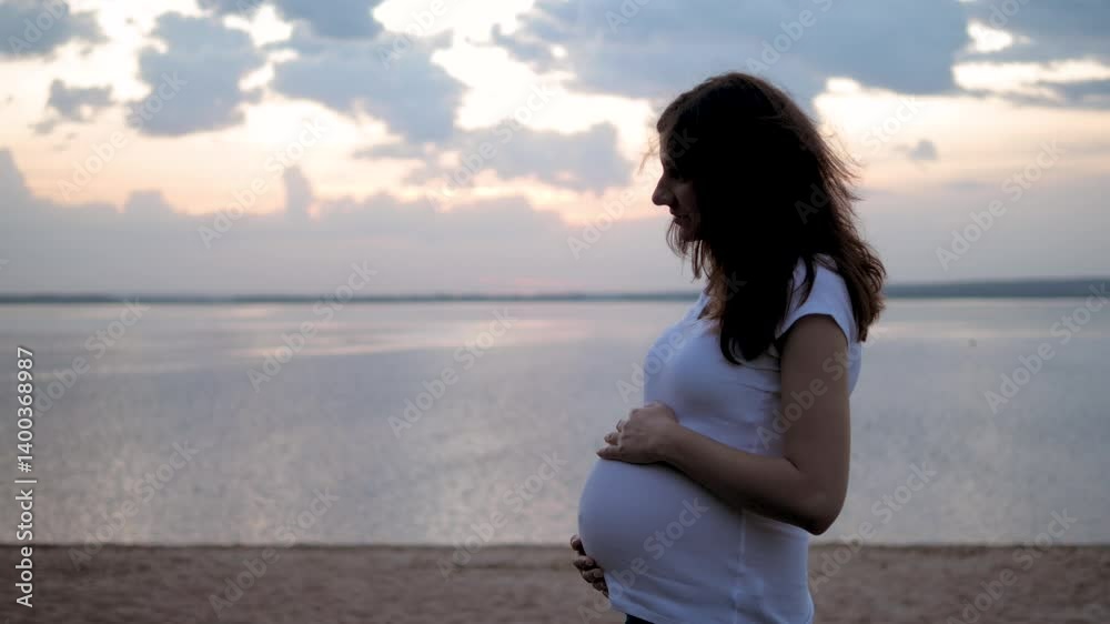 Gentle sunlight illuminates a pregnant woman standing by a tranquil lake shore. Hands lovingly stroked her baby bump as she enjoys serene sunset view. Gentle sunlight illuminates a pregnant woman standing by a tranquil lake shore. Hands lovingly stroked her baby bump as she enjoys serene sunset view.