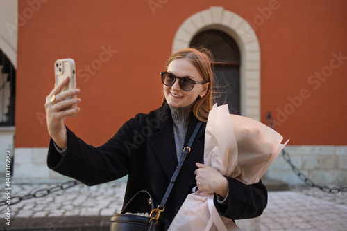 A young woman with sunglasses and a black coat taking a selfie while holding a bouquet of flowers. She appears happy and is posing in front of a colorful building, capturing a joyful moment in the
