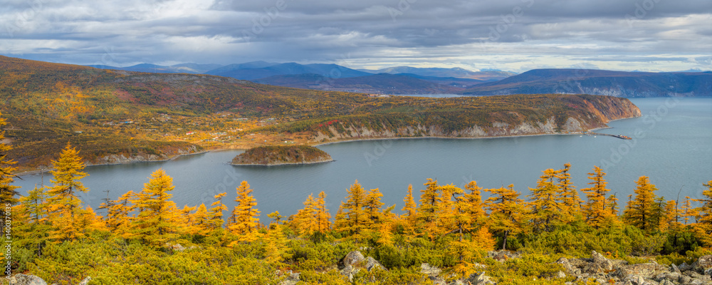 Beautiful autumn panorama. View from the hill to the sea bay, cape and island. Dwarf pines and larch trees on a mountain slope. Village on the coast. Mountains in the distance. Magadan Region, Russia.