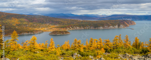 Obraz na płótnie Beautiful autumn panorama. View from the hill to the sea bay, cape and island. Dwarf pines and larch trees on a mountain slope. Village on the coast. Mountains in the distance. Magadan Region, Russia. w salonie