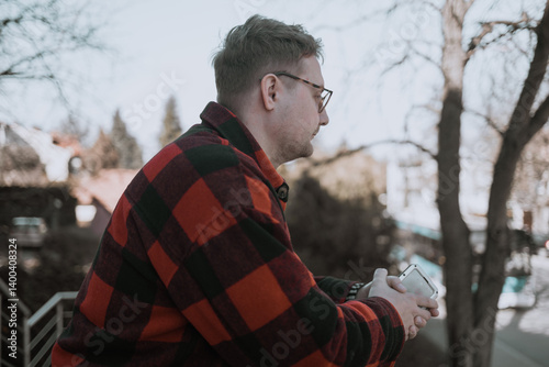 A young blond man wearing glasses, looking at his phone while walking down the street. A modern scene showcasing urban life, technology, and casual street style