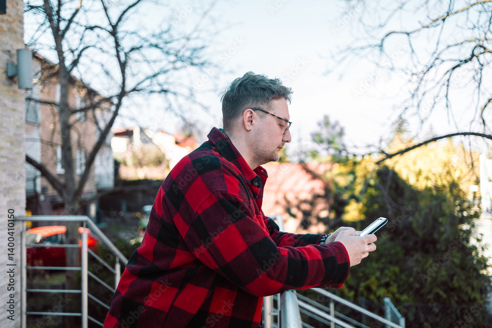 A young blond man wearing glasses, looking at his phone while walking down the street. A modern scene showcasing urban life, technology, and casual street style