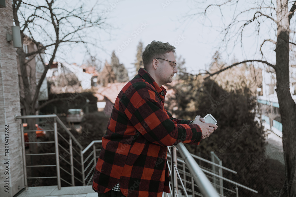A young blond man wearing glasses, looking at his phone while walking down the street. A modern scene showcasing urban life, technology, and casual street style
