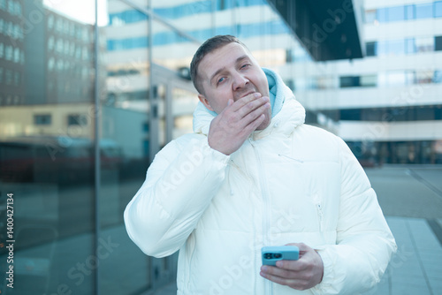 Caucasian 30s man using smartphone outdoors in winter bored yawning tired covering mouth with hand. restless and sleepiness. High quality photo