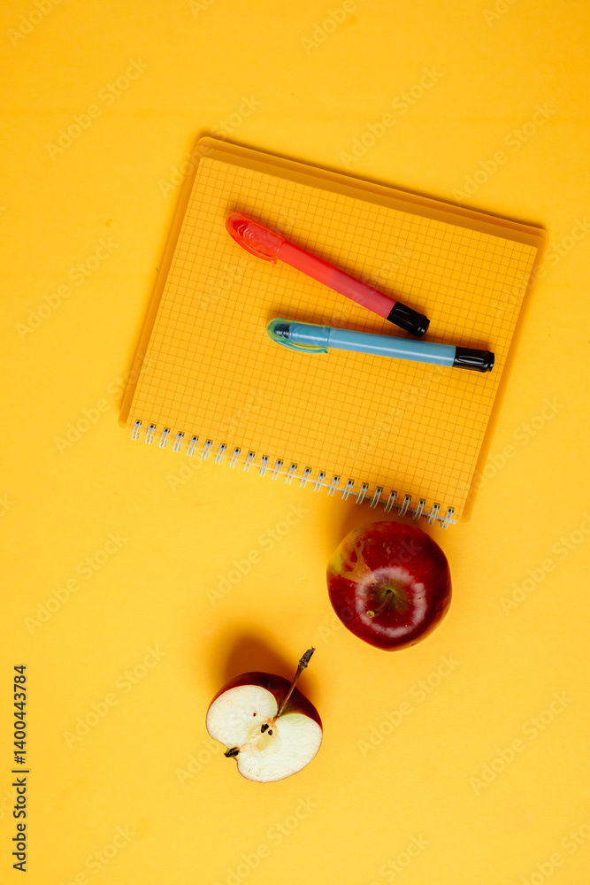 A vibrant and colorful setup featuring office supplies such as pens, sticky notes, and a notepad, complemented by a fresh red apple. The bright yellow background adds an energetic touch to the scene