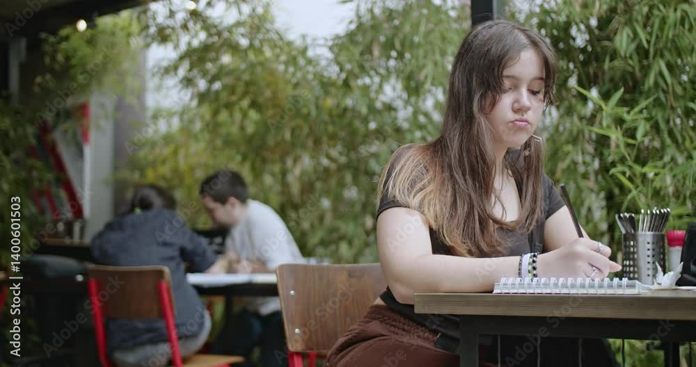 Young Female College Student Studying and Taking Notes in Notebook at Outdoor Cafe Table With Bamboo and Greenery in Background