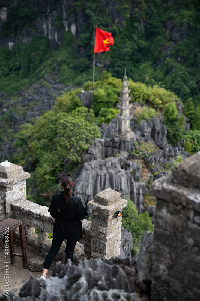 (Selective focus) Stunning view of a girl enjoying the view from the Hang Múa Viewpoint with a pagoda and the Vietnamese national flag in the distance. Ninh Binh, Vietnam.