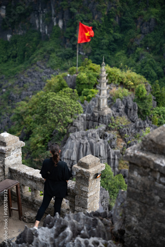 Obraz na płótnie (Selective focus) Stunning view of a girl enjoying the view from the Hang Múa Viewpoint with a pagoda and the Vietnamese national flag in the distance. Ninh Binh, Vietnam. w salonie