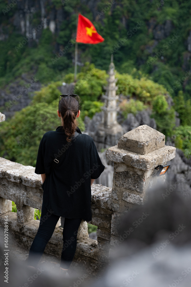 (Selective focus) Stunning view of a girl enjoying the view from the Hang Múa Viewpoint with a pagoda and the Vietnamese national flag in the distance. Ninh Binh, Vietnam.