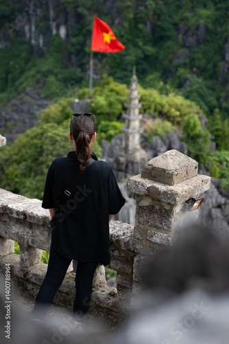 Obraz na płótnie (Selective focus) Stunning view of a girl enjoying the view from the Hang Múa Viewpoint with a pagoda and the Vietnamese national flag in the distance. Ninh Binh, Vietnam. w salonie