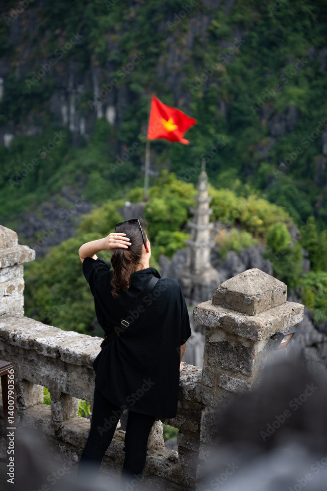 (Selective focus) Stunning view of a girl enjoying the view from the Hang Múa Viewpoint with a pagoda and the Vietnamese national flag in the distance. Ninh Binh, Vietnam.