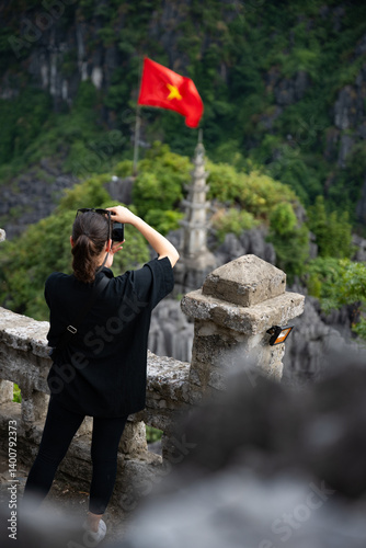 Obraz na płótnie (Selective focus) A girl is taking pictures from the Hang Múa Viewpoint with a pagoda and the Vietnamese national flag in the distance. Ninh Binh, Vietnam. w salonie
