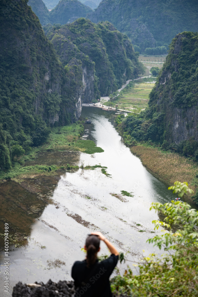(Selective focus) Defocused girl taking pictures from the Hang Múa Viewpoint with the a stunning landscape and the Tam Coc river in the distance. Ninh Binh, Vietnam.