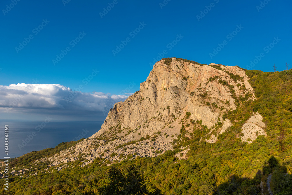 A monumental rock on the southern coast of Crimea is illuminated by bright sunlight against a clear blue sky. In the foreground is a dense green forest, A monumental rock on the southern coast of Crimea is illuminated by bright sunlight against a clear blue sky. In the foreground is a dense green forest,
