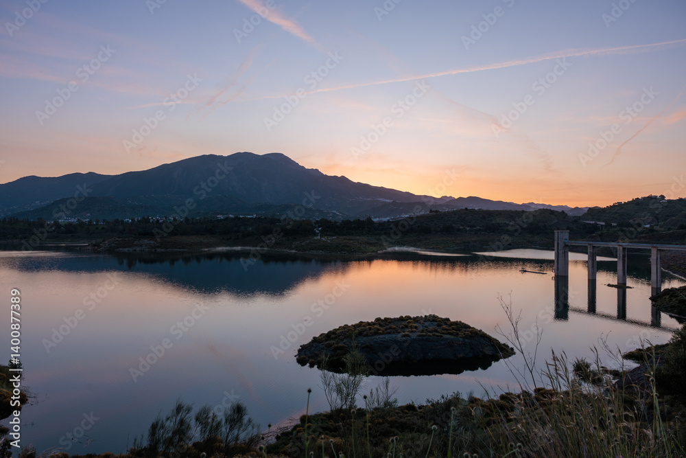 Calm lake reflecting mountain range at sunset with concrete bridge structure