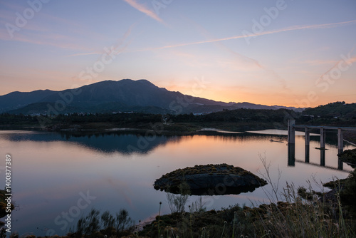 Obraz na płótnie Calm lake reflecting mountain range at sunset with concrete bridge structure w salonie