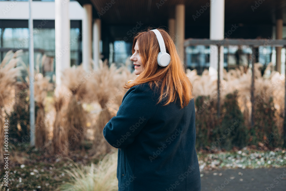 Beautiful young happy woman in headphones with bouquet of tulip posing on urban background. Smiling 30s girl listening to music through wireless headphones, rear view