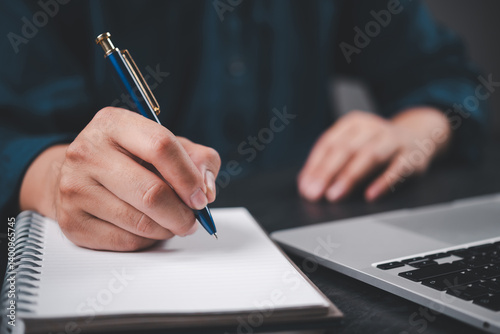 Close-up hand writing on notebook with pen next to laptop. Education, business planning, journaling setup with modern workspace productivity and creative thinking.