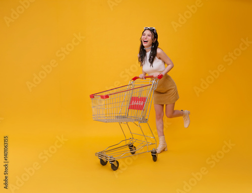 Full length happy excited Asian teen woman pushing empty shopping cart or shopping trolley grocery running promotion supermarket isolated on yellow background.