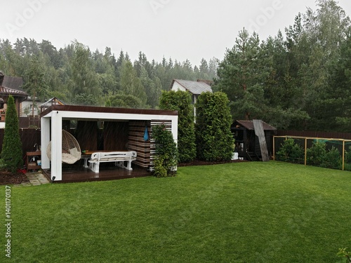 A wooden gazebo with benches, table and swings stands next to a mowed grassy lawn. Behind it are pine, birch and thuja trees, a metal fence and a forest. Cloudy summer weather, wind and heavy rain