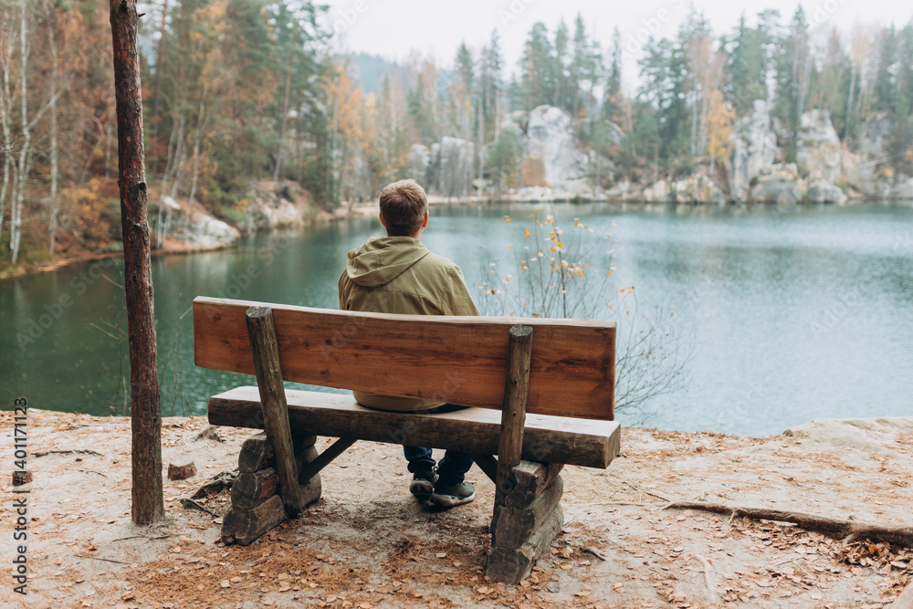 Lonely 30s blonde man is sitting on the bench of a forest lake. Autumn landscape, calmness, tranquility, travel concept. A man on the bank of a lake is enjoying the nature. High quality photo.