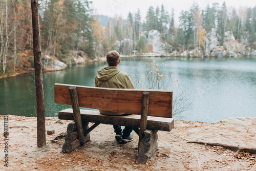 Lonely 30s blonde man is sitting on the bench of a forest lake. Autumn landscape, calmness, tranquility, travel concept. A man on the bank of a lake is enjoying the nature. High quality photo.