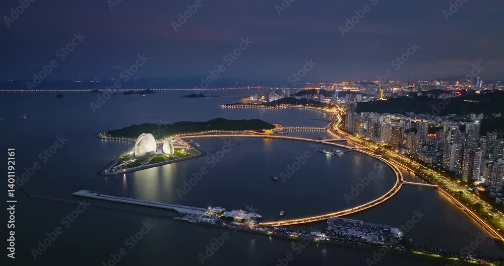 Aerial view of the illuminated Zhuhai city skyline and surrounding cityscape with coastal bridge lights reflecting on the water at night.