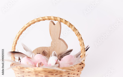 Dekoracja, wiklinowy koszyk wielkanocny z pastelowymi jajkami, w kolorach białym i różowym, z piórkami i drewnianym ozdobnym zającem w  tle photo