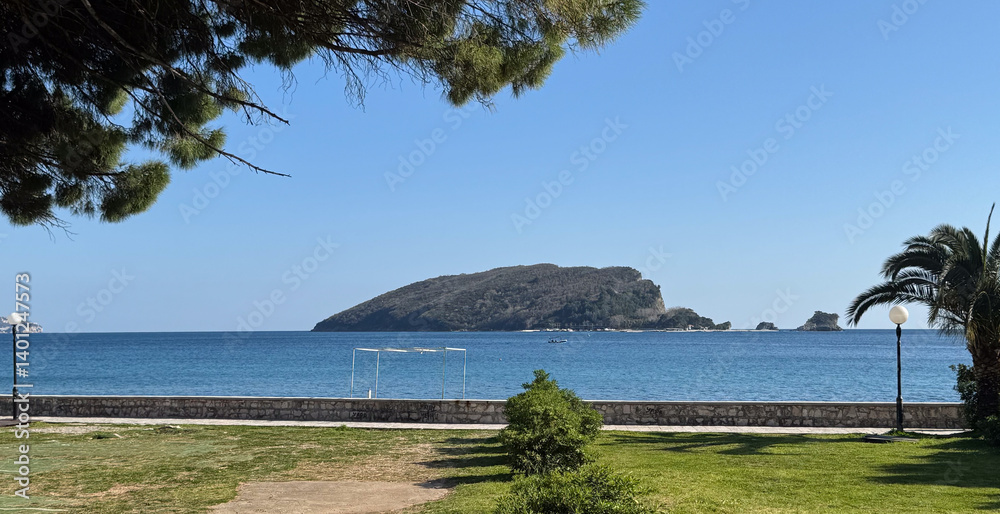 Scenic view of forested island surrounded by tranquil blue sea, framed by palm trees and coastal park.