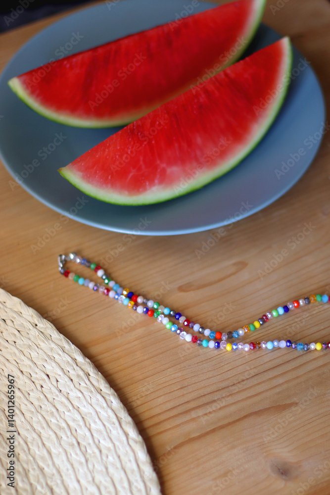 Plate with watermelon slices, glass of orange juice or cocktail, straw hat, colorful beaded necklace and sunglasses on the wooden table. Summer essentials concept. Selective focus.