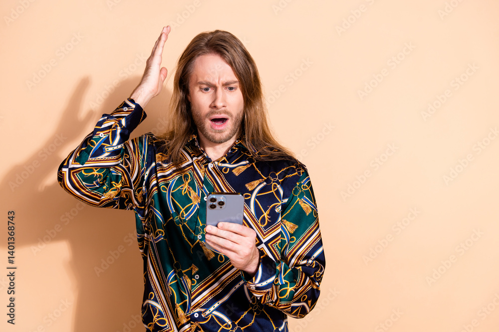 Concerned young man in stylish printed shirt holding smartphone, beige background
