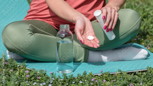 Young woman holding soluble tablet in hand, making effervescent sports drink, sitting on yoga mat during outdoor sport exercise. Nutrition supplements or prevention of vitamin deficiency