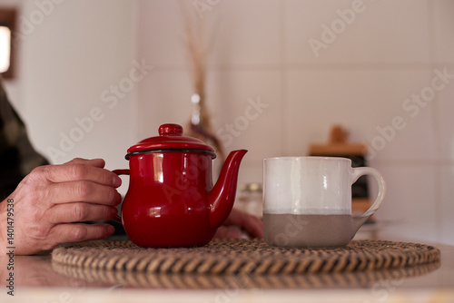 Elderly man gently holding a vintage red teapot, preparing a comforting morning tea ritual, with a ceramic mug placed nearby on a woven mat in a cozy kitchen setting