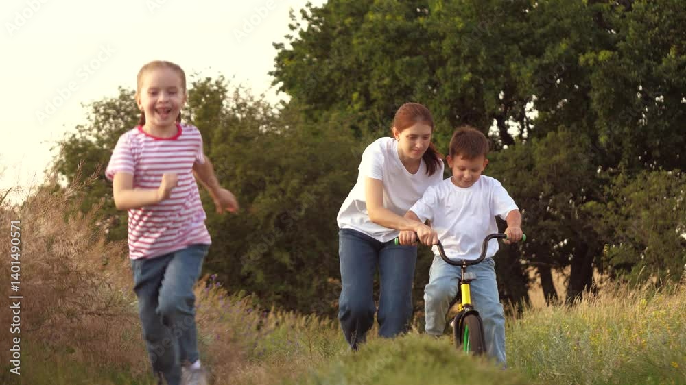 Smiling girl runs while mother teaches little boy to ride bicycle on grassy rural road at sunset. Mother with girl and boy practice bicycle riding in field. Caring mother with boy and girl on vacation Smiling girl runs while mother teaches little boy to ride bicycle on grassy rural road at sunset. Mother with girl and boy practice bicycle riding in field. Caring mother with boy and girl on vacation