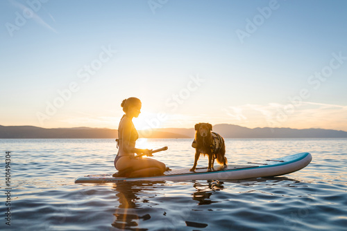 Girl with a dog sitting on a sup board surfing on a lake in a sunset. Dog is wearing a life vest and girl is wearing a swimsuit