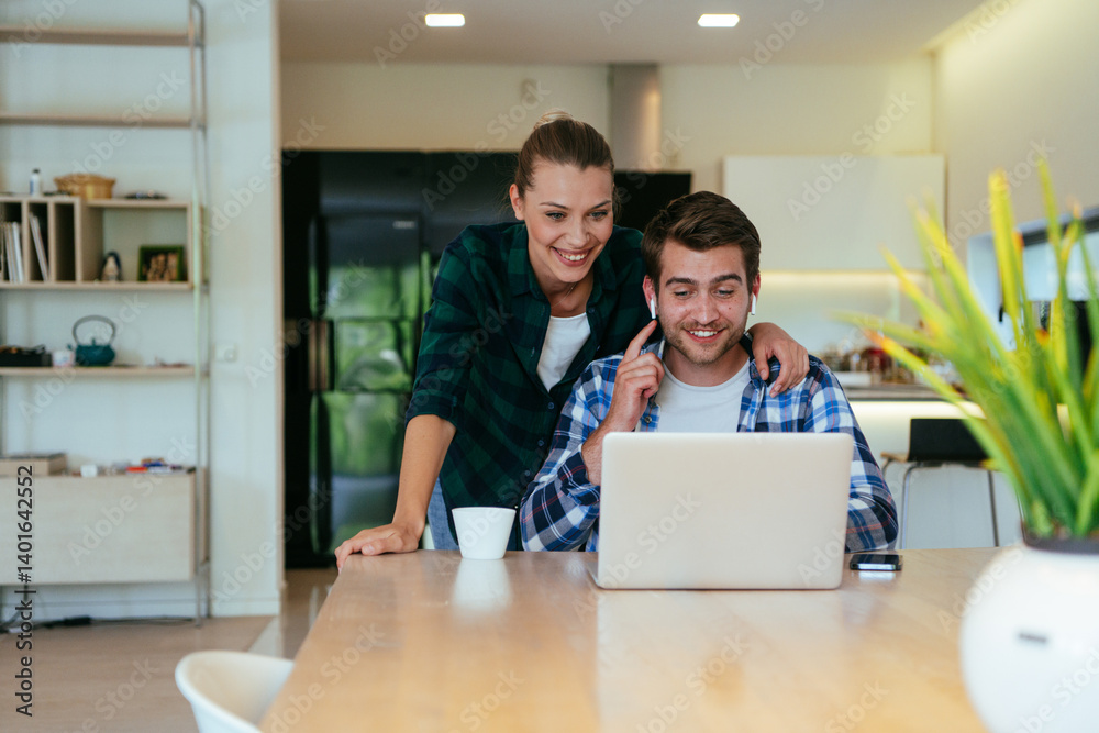 A young married couple is talking to parents, family and friends on a video call via a laptop while sitting in the living room of their modern house.